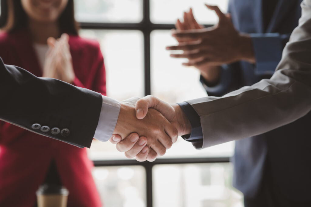Two business professionals shaking hands, with their colleagues clapping hands in the background.