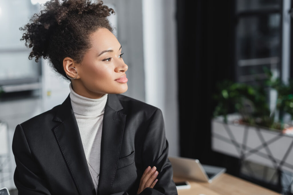 A young professional with her arms crossed, looking outside a modern office setting.